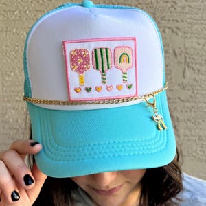 May include: A trucker hat with a white front panel and a light blue brim and mesh back. The front features a patch with three pickleball paddles in pink, green, and orange, with gold chain and a pickleball charm.