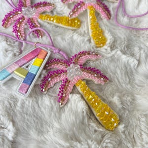 Op de afbeelding: Beaded palmboom hangers in roze, geel en heldere kristallen, met een regenboogkleurige letter "A" hanger. De hangers zijn geregen aan dunne, lichtpaarse koorden, tegen een pluizige witte achtergrond.