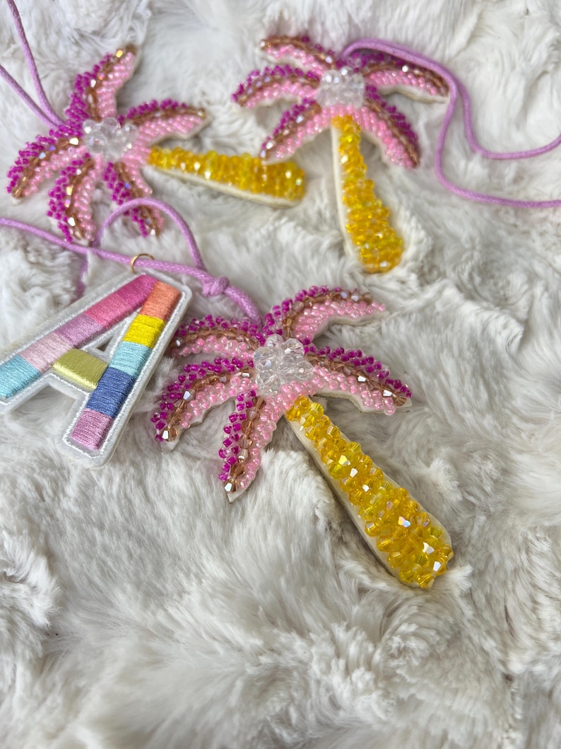 Op de afbeelding: Beaded palmboom hangers in roze, geel en heldere kristallen, met een regenboogkleurige letter "A" hanger. De hangers zijn geregen aan dunne, lichtpaarse koorden, tegen een pluizige witte achtergrond.
