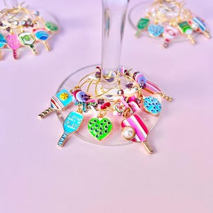 May include: Wine glass charms with colorful pickleball paddle and heart designs. Charms are attached to gold-colored wire rings. The paddles are in various colors, including blue, pink, and green, with some featuring the text "LET'S PLAY".