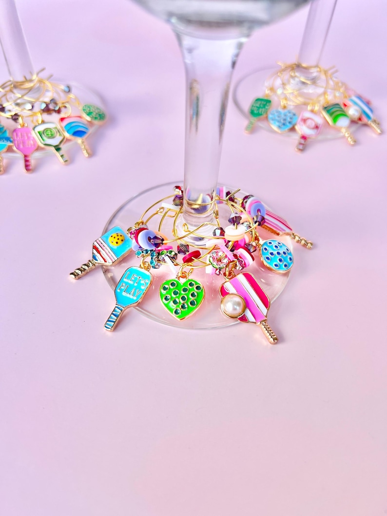 May include: Wine glass charms with colorful pickleball paddle and heart designs. Charms are attached to gold-colored wire rings. The paddles are in various colors, including blue, pink, and green, with some featuring the text "LET'S PLAY".