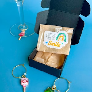 May include: A black gift box with a small package that has a sticker that says "I hope this makes you smile". Two wine charms with pickleball paddle designs are on a blue background. One wine glass has a charm.