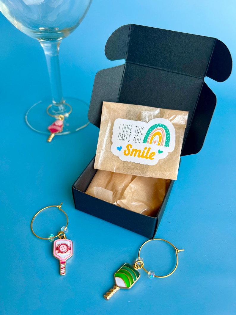 May include: A black gift box with a small package that has a sticker that says "I hope this makes you smile". Two wine charms with pickleball paddle designs are on a blue background. One wine glass has a charm.