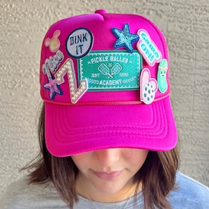 May include: A bright pink trucker hat with a variety of colorful patches, including a green patch that says "Pickle Baller Academy est. 1985", a blue patch that says "Come On", and a white patch that says "Dunk It".