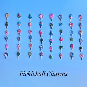 May include: A collection of colorful pickleball charm pendants on a light blue background. The charms feature various designs, including stripes, hearts, and geometric patterns. The text "Pickleball Charms" is displayed at the bottom.