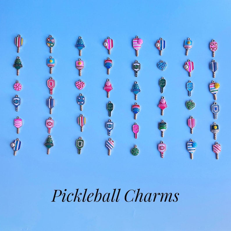 May include: A collection of colorful pickleball charm pendants on a light blue background. The charms feature various designs, including stripes, hearts, and geometric patterns. The text "Pickleball Charms" is displayed at the bottom.