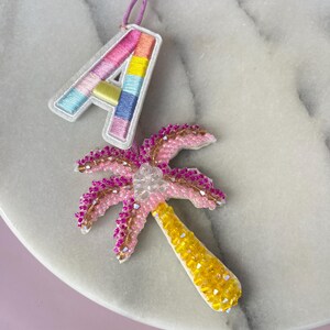 Op de afbeelding: Een close-up van twee decoratieve items: een kleurrijk geborduurde letter "A" en een kralenpalmboom. De letter "A" is wit omlijnd en heeft regenboogkleurige strepen. De palmboom is roze met een gele stam en heldere kralen.