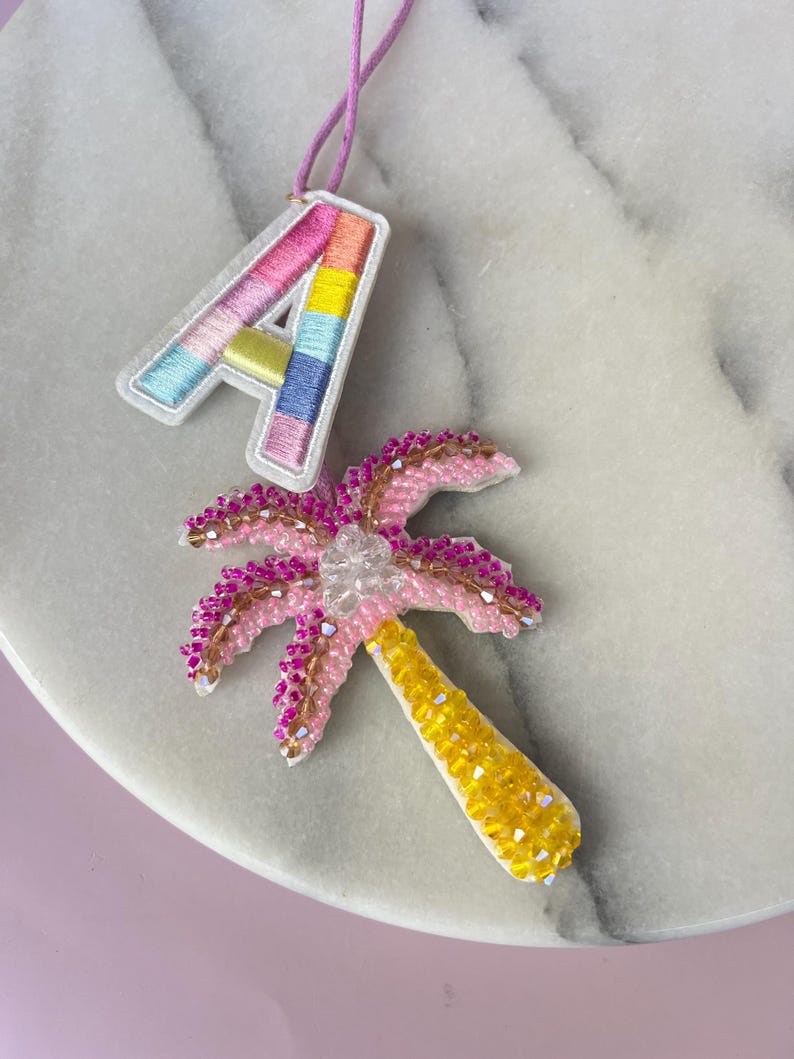 Op de afbeelding: Een close-up van twee decoratieve items: een kleurrijk geborduurde letter "A" en een kralenpalmboom. De letter "A" is wit omlijnd en heeft regenboogkleurige strepen. De palmboom is roze met een gele stam en heldere kralen.