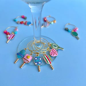 May include: A collection of colorful pickleball-themed wine charms. Each charm features a miniature pickleball paddle design with various patterns and colors, including blue, pink, green, and rainbow stripes. The charms are attached to gold-colored hoops.