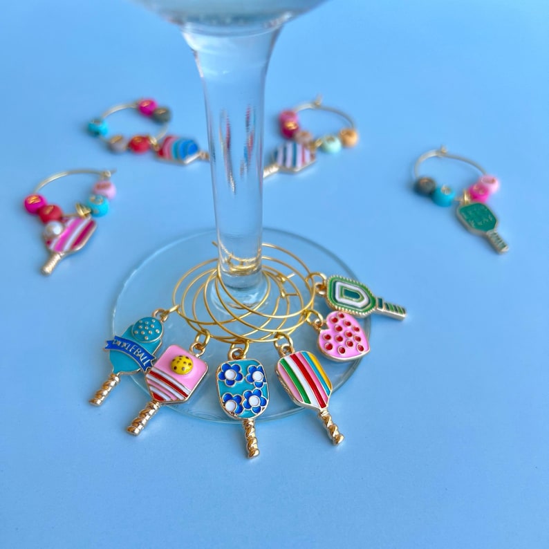 May include: A collection of colorful pickleball-themed wine charms. Each charm features a miniature pickleball paddle design with various patterns and colors, including blue, pink, green, and rainbow stripes. The charms are attached to gold-colored hoops.