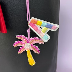 Op de afbeelding: Een close-up van een roze koord met twee geborduurde patches. E&eacute;n patch is een kleurrijke letter "A" met een witte rand. De andere is een roze palmboom met een gele stam en witte accenten. De achtergrond is zwart.