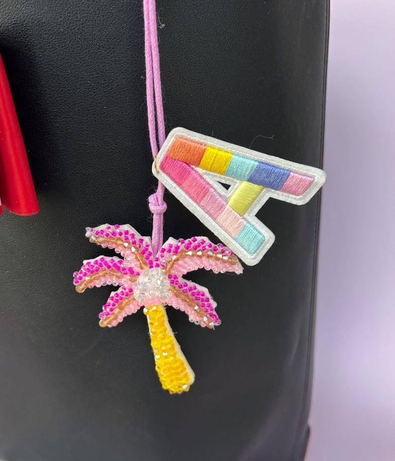 Op de afbeelding: Een close-up van een roze koord met twee geborduurde patches. E&eacute;n patch is een kleurrijke letter "A" met een witte rand. De andere is een roze palmboom met een gele stam en witte accenten. De achtergrond is zwart.