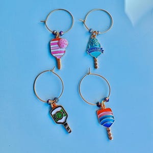 May include: Four gold-colored wine charms with colorful pickleball paddle designs. Each charm has a different paddle design in shades of pink, blue, green, and orange. The charms are attached to gold hoops with decorative beads.
