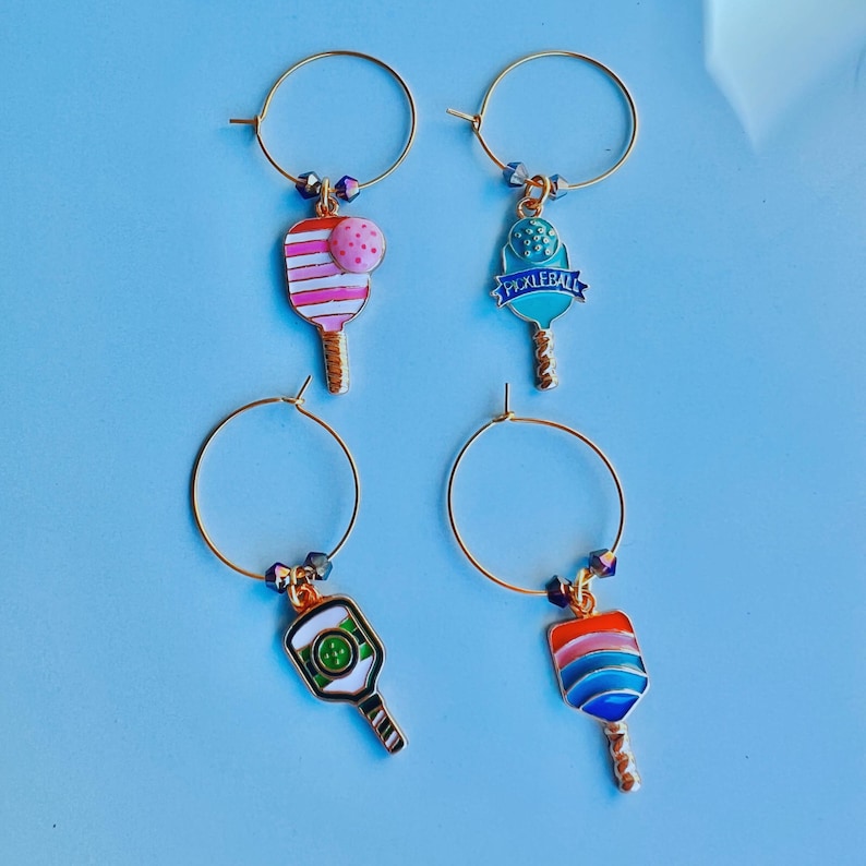May include: Four gold-colored wine charms with colorful pickleball paddle designs. Each charm has a different paddle design in shades of pink, blue, green, and orange. The charms are attached to gold hoops with decorative beads.