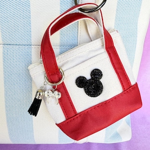 May include: A miniature white canvas tote bag keychain with red trim and handles. The front features a black rhinestone mouse ear silhouette. A silver metal ring attaches a small black tassel and a silver-toned bow charm to the bag handle.