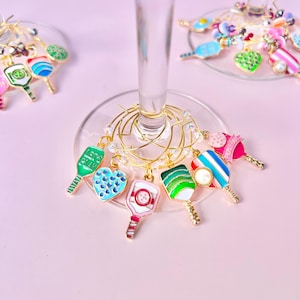 May include: A collection of colorful wine charms, each featuring a unique design. Charms include a green paddle, a blue heart, and a pink and white striped paddle. The charms are attached to gold wire rings, perfect for identifying wine glasses.
