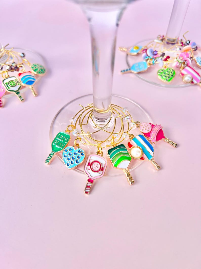 May include: A collection of colorful wine charms, each featuring a unique design. Charms include a green paddle, a blue heart, and a pink and white striped paddle. The charms are attached to gold wire rings, perfect for identifying wine glasses.