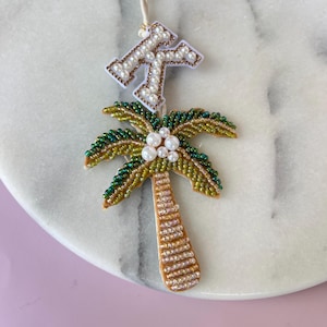 Op de afbeelding: Een met kralen versierd palmboomornament met een witte en gouden initiaal. De palmboom heeft groene en gouden bladeren, een bruine stam en witte parelaccenten. De initiaal is wit met gouden en parelaccenten.