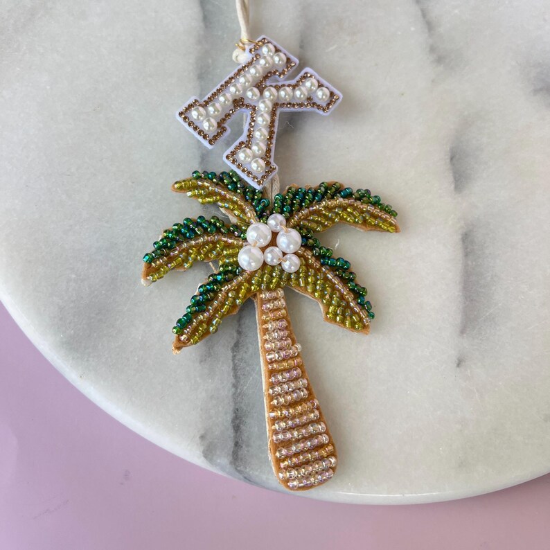 Op de afbeelding: Een met kralen versierd palmboomornament met een witte en gouden initiaal. De palmboom heeft groene en gouden bladeren, een bruine stam en witte parelaccenten. De initiaal is wit met gouden en parelaccenten.