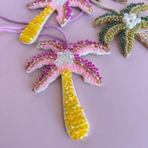 Op de afbeelding: Handgemaakte palmboomhangers van kralen in roze, geel en groen. De hangers hebben ingewikkeld kralenwerk, met een witte basis en een helder kristallen accent aan de bovenkant. Elke hanger is geregen aan een dun koord.