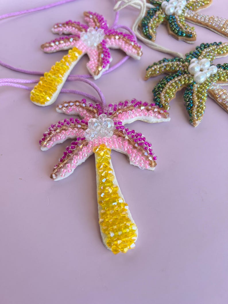 Op de afbeelding: Handgemaakte palmboomhangers van kralen in roze, geel en groen. De hangers hebben ingewikkeld kralenwerk, met een witte basis en een helder kristallen accent aan de bovenkant. Elke hanger is geregen aan een dun koord.