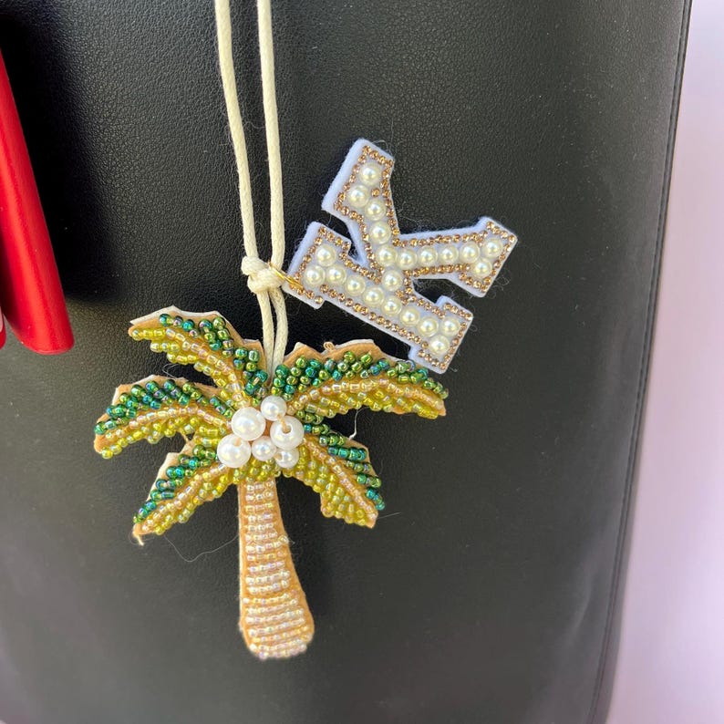 Op de afbeelding: Een kralen palmboom en letter "K" bedel hangen aan een cr&egrave;mekleurig touwtje. De palmboom heeft groene en gele kralen met witte parelaccenten. De letter "K" is wit met parel- en gouden kraalaccenten.