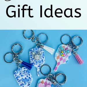 May include: A variety of pickleball-themed gift ideas are displayed. The image includes a white blanket with colorful pickleball graphics, keychains with decorative charms, and wine glass charms. The text "Pickleball Gift Ideas" is also visible.