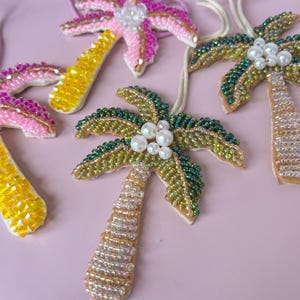 Op de afbeelding: Close-up van verschillende kralen palmboom hangers. De hangers hebben kleurrijke kralen in roze, geel, groen en goud. Elke hanger heeft een wit touwtje om op te hangen en is versierd met imitatieparels.