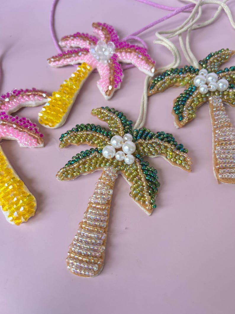 Op de afbeelding: Close-up van verschillende kralen palmboom hangers. De hangers hebben kleurrijke kralen in roze, geel, groen en goud. Elke hanger heeft een wit touwtje om op te hangen en is versierd met imitatieparels.