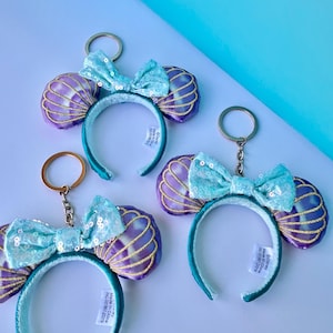 May include: Three keychains featuring teal headbands with seashell-shaped ears in purple and gold. Each keychain has a teal sequined bow. The background is a gradient of white and light blue.