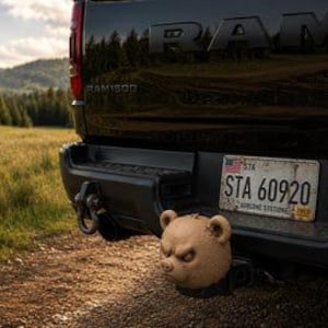 May include: A black RAM 1500 truck rear bumper featuring a tan bear head hitch cover. The vehicle has a license plate reading STA 60920 with the text AIRBORNE STATION. The hitch cover is shaped like a bear face with visible ears and eyes.