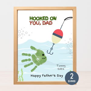May include: A framed print with the text "HOOKED ON YOU, DAD" in green and red. The artwork features a handprint fish with a fishing bobber and hook. The text "Happy Father's Day" is also included.