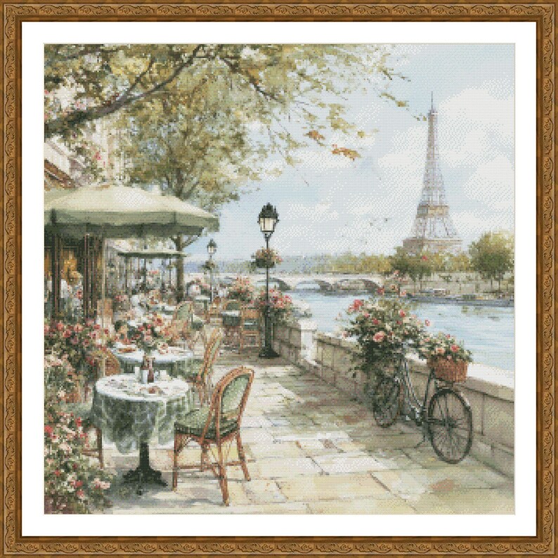 May include: A framed cross-stitch depicting a Parisian cafe scene. The image features tables with green tablecloths, chairs, and a bicycle with a basket of flowers. The Eiffel Tower is visible in the background, along with a river and bridge.