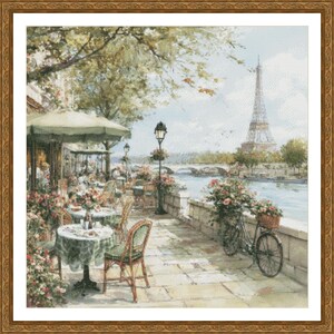 May include: A framed cross-stitch depicting a Parisian cafe scene. The image features tables with green tablecloths, chairs, and floral arrangements. The Eiffel Tower is visible in the background, along with a bicycle with a basket.
