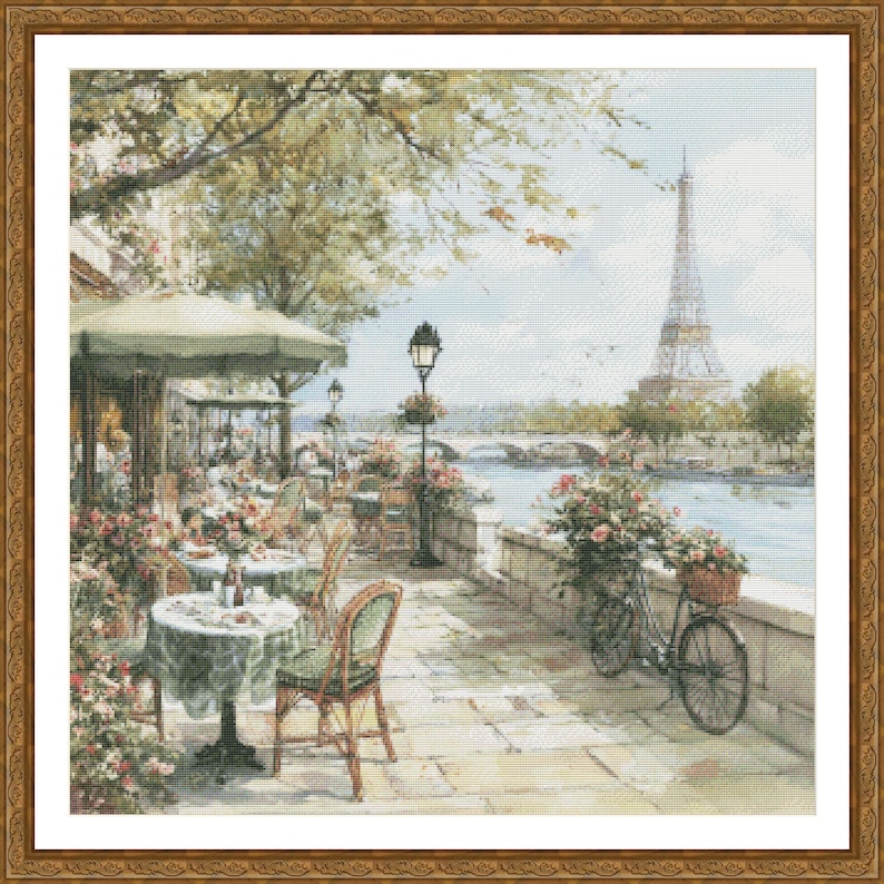 May include: A framed cross-stitch depicting a Parisian cafe scene. The image features tables with green tablecloths, chairs, and a bicycle with a basket of flowers. The Eiffel Tower is visible in the background, along with a river and bridge.