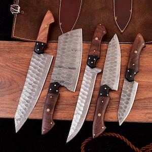 May include: A set of six kitchen knives with wooden handles and patterned steel blades. The handles are a dark brown color with brass rivets. The blades have a unique, textured pattern. The knives are arranged on a wooden surface.