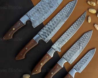 May include: An overhead shot of a set of five Damascus steel kitchen knives with wooden handles. The blades have a unique patterned design. The handles are a dark brown color with brass rivets. The knives are arranged on a brown and black surface.