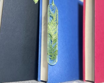 May include: A clear bookmark with a pressed green leaf inside. The bookmark is attached to a blue book with a green tassel.