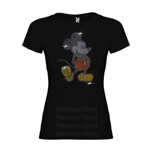 Può includere: Maglietta nera con un design di Mickey Mouse in strass. Il design presenta Mickey Mouse in un abito rosso e giallo con un contorno nero. Il testo "Rhinestone Street" è ripetuto quattro volte nella parte inferiore della maglietta.