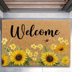 May include: A rectangular doormat with a brown background and the word "Welcome" in black script. The mat features a floral design with yellow sunflowers, daffodils, and a butterfly. The doormat is placed in a doorway.