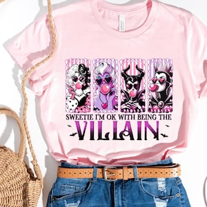 May include: A light pink t-shirt with a graphic of four cartoon villains blowing bubblegum bubbles. The text reads "Sweetie I'm ok with being the Villain". The shirt is paired with denim shorts, a straw bag, and sandals.