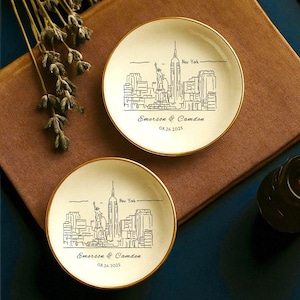 May include: Two small, off-white plates with gold rims, each featuring a black line drawing of the New York City skyline, including the Empire State Building and the Statue of Liberty. The plates are personalized with the names "Emerson & Camden" and the date "08.26.2025".