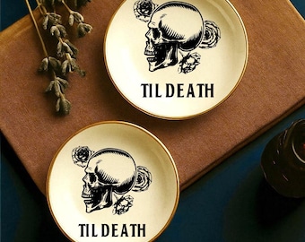 May include: Two small, round, cream-colored plates with gold rims, each featuring a black skull design with roses and the text "TIL DEATH." The plates rest on a brown surface, with a dark blue background and decorative elements.