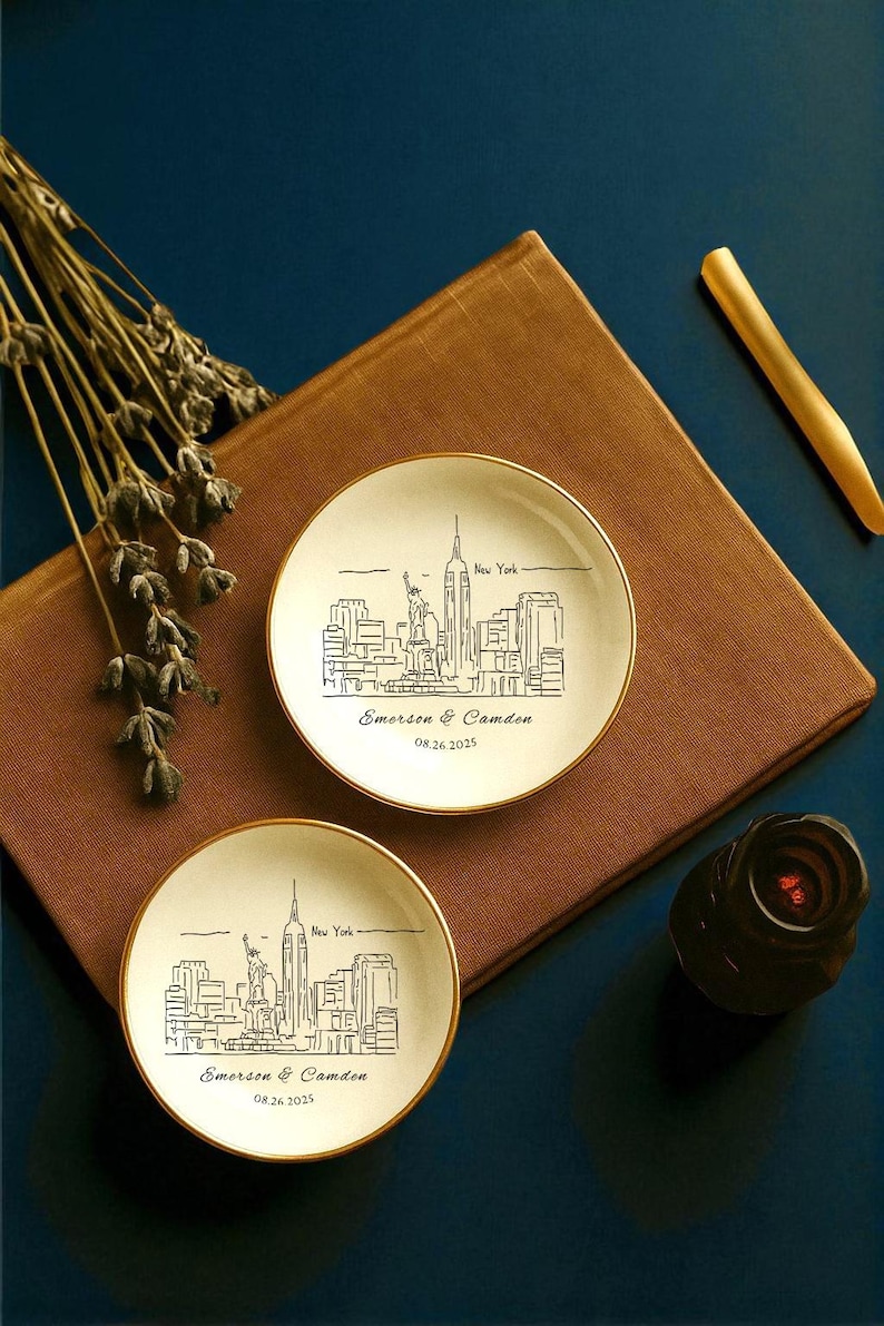 May include: Two small, off-white plates with gold rims, each featuring a black line drawing of the New York City skyline, including the Empire State Building and the Statue of Liberty. The plates are personalized with the names "Emerson & Camden" and the date "08.26.2025".