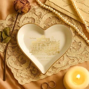May include: Heart-shaped, white ceramic trinket dish with a gold rim and a gold illustration of a building. The dish has text that reads "From Rome" and "Sergio & Carina" with a date. A lit candle, rings, and a pearl necklace are nearby.