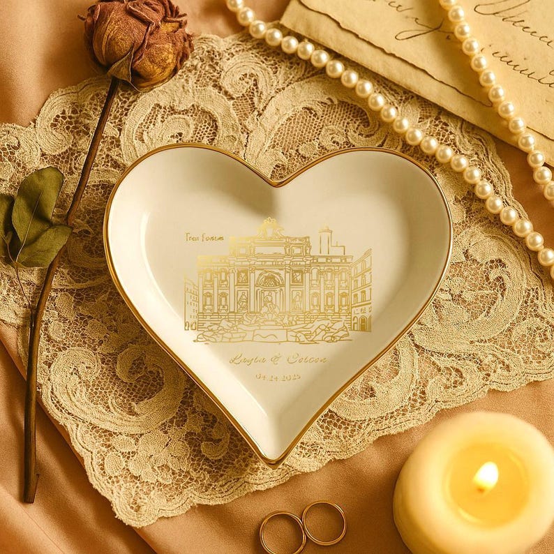 May include: Heart-shaped, white ceramic trinket dish with a gold rim and a gold illustration of a building. The dish has text that reads "From Rome" and "Sergio & Carina" with a date. A lit candle, rings, and a pearl necklace are nearby.