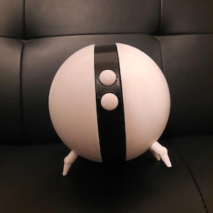 May include: A white, spherical object with a black vertical stripe and two white circular buttons. The object has three small, white legs. The design is minimalist and modern, with a clean aesthetic.