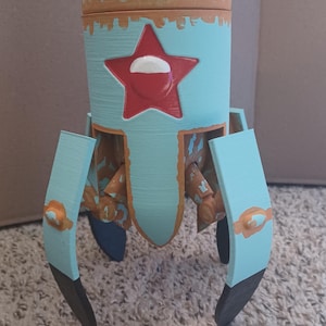 Może przedstawiać: Ręcznie malowany, jasnoniebieski pojemnik w kształcie rakiety z czerwoną gwiazdą i białym akcentem. Pojemnik ma brązowe wykończenia i nogi z czarnymi końcówkami. Rakieta jest przeznaczona do przechowywania drobnych przedmiotów.