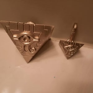 May include: Two silver-coloured triangular pendants. One is a ring, the other a pendant with a loop. Both feature an eye design within the triangle. Likely for a necklace or keyring.