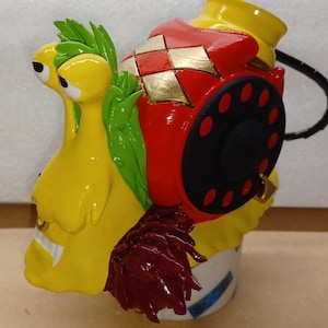 Puede incluir: Figura de caracol de cerámica pintada a mano. El caracol es principalmente amarillo con hojas verdes, una concha roja con detalles dorados y un dial de teléfono negro. La figura tiene un diseño de dibujos animados.